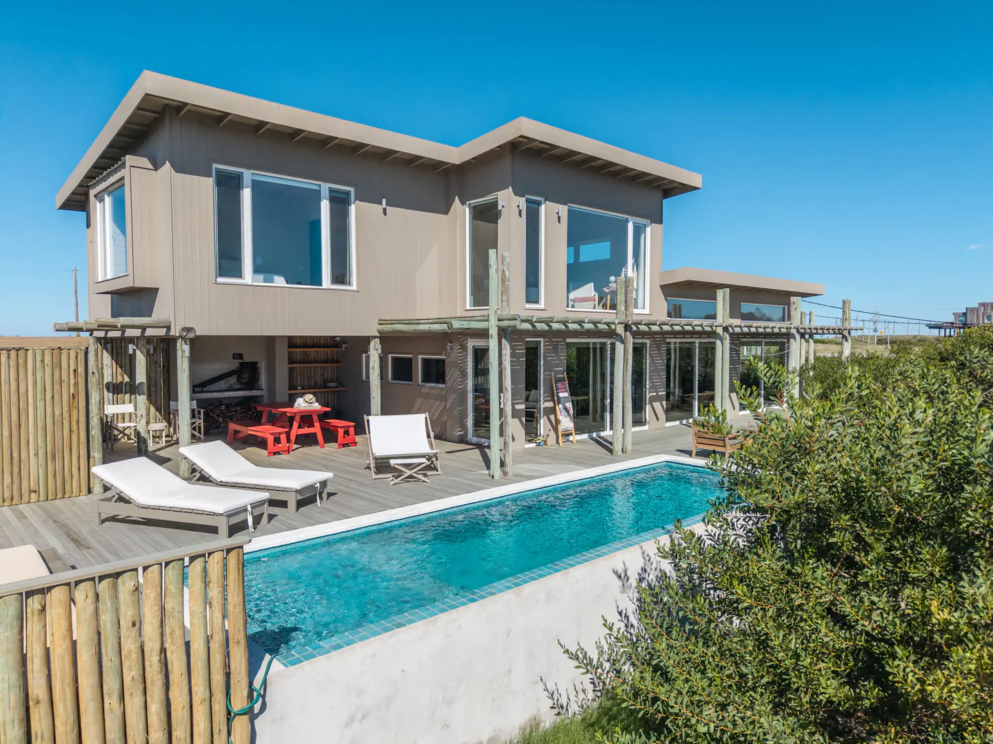 Two-story beige house with a pool, deck, and outdoor furniture under a clear blue sky.