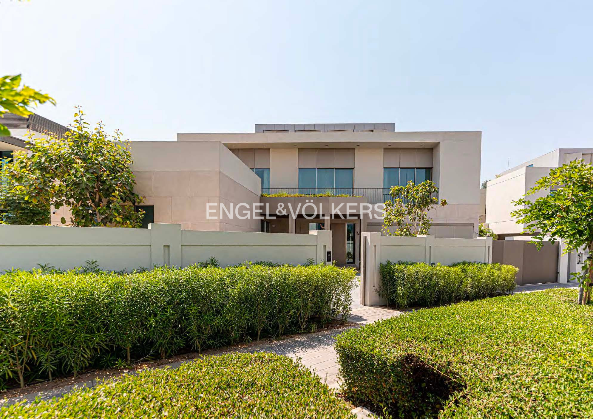 Modern two-story beige villa with large windows, surrounded by manicured green hedges and a stone wall under a clear sky. [AI-generated text]