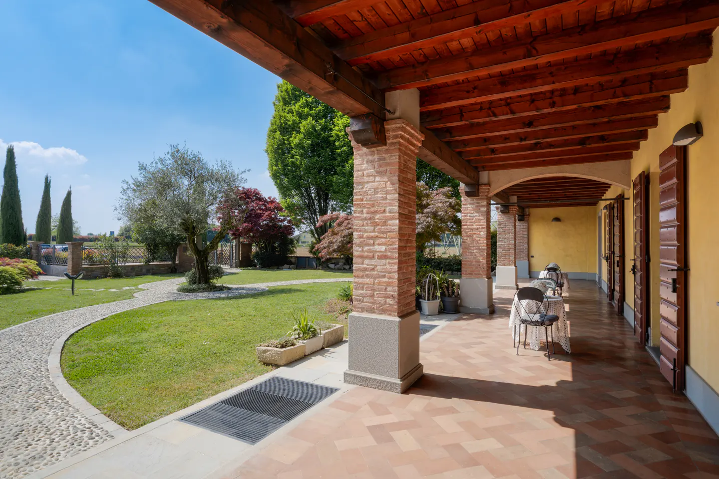 Covered patio with brick columns and wood ceiling overlooks a green lawn and garden on a sunny day.