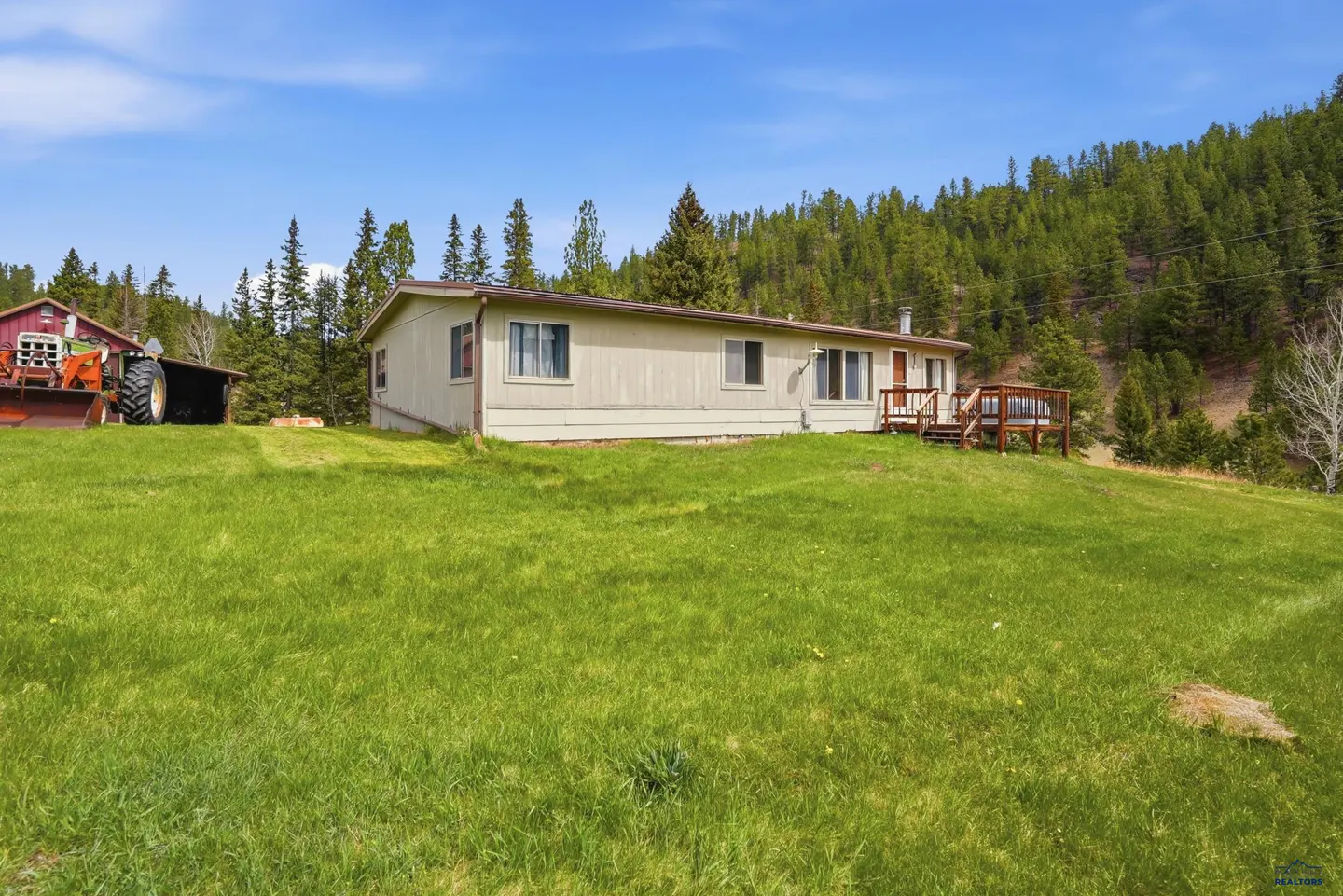 A single-story beige home with a wooden deck sits on a grassy hill near a forest, with a tractor in a nearby shed.