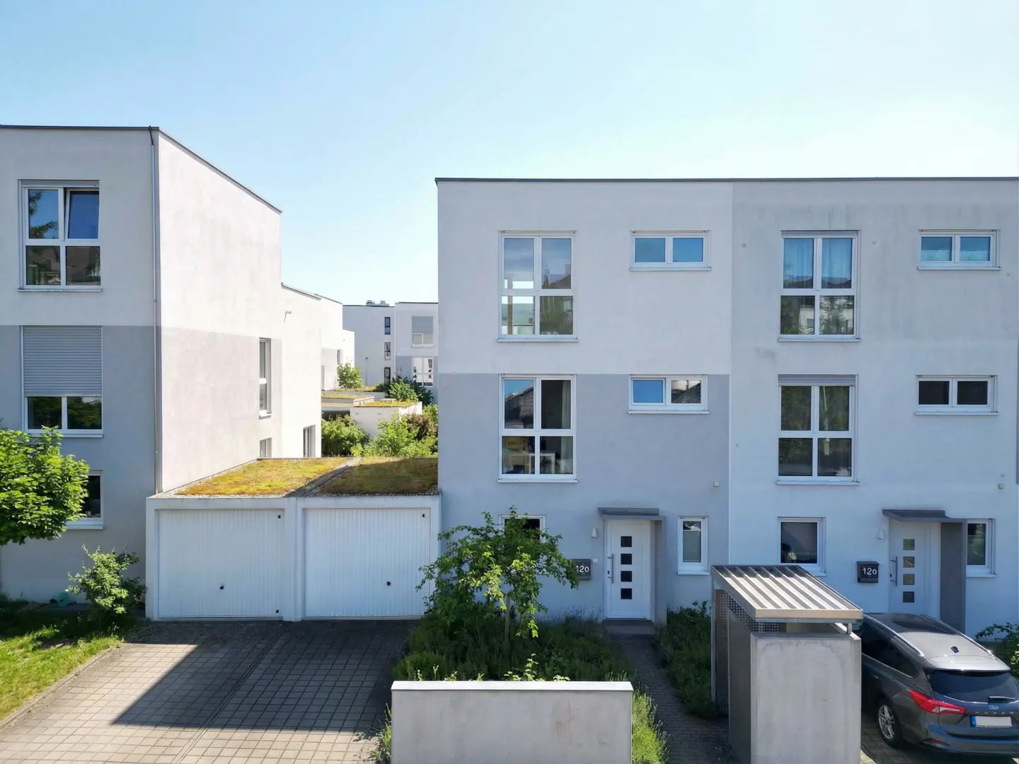 Modern townhouses with white and gray exteriors, large windows, and green roofs on garages. A car is parked in front.