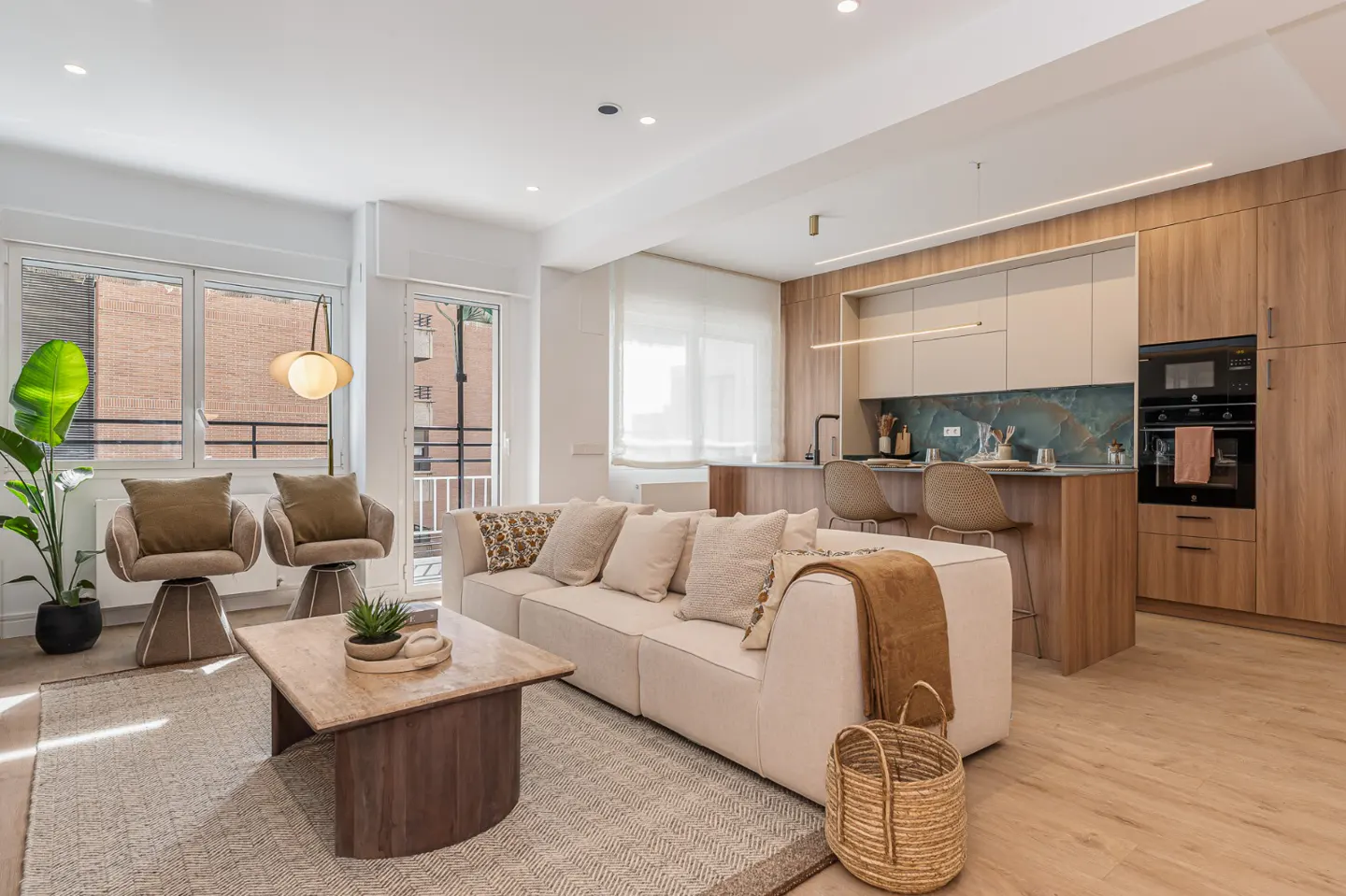 Bright, modern living room with a beige sofa, two chairs, and an open kitchen with wood cabinets. A woven basket sits on the light wood floor.