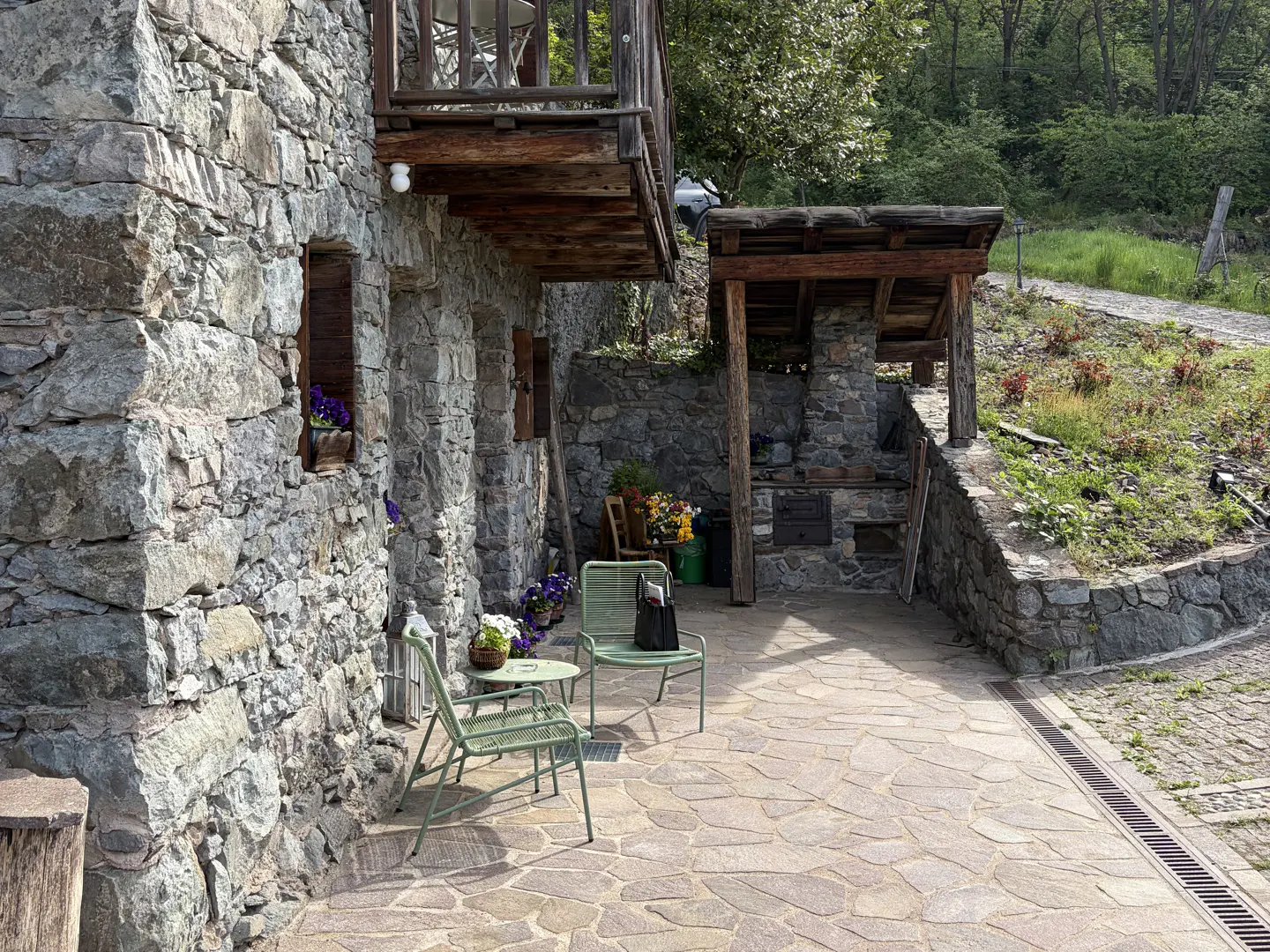 Stone house exterior with a patio featuring green metal chairs and table, stone oven, and a balcony above.