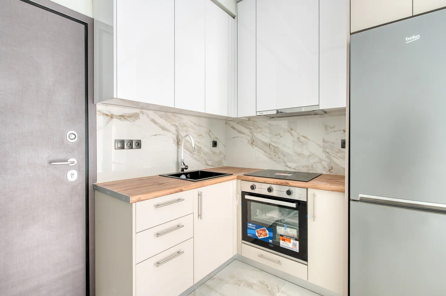 A compact kitchen features white cabinets, a wood-look countertop, and a stainless steel refrigerator. A gray door is visible on the left.