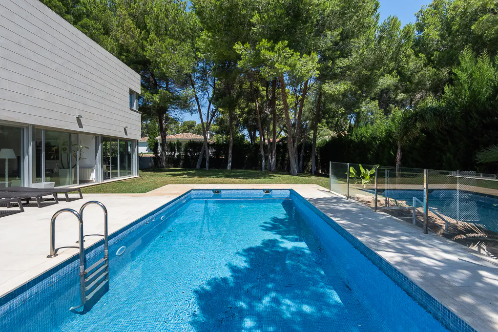 A backyard with a rectangular blue tiled pool, a ladder, and a modern house with large windows. Trees are in the background.