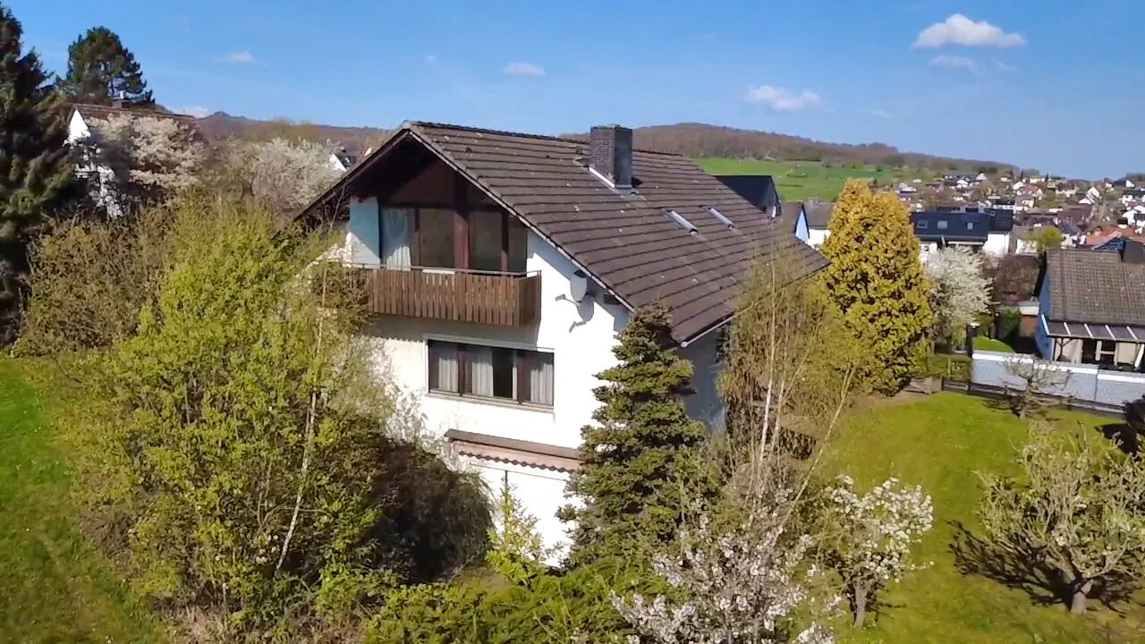 Two-story white house with brown roof and balcony, surrounded by green trees and grass on a sunny day.