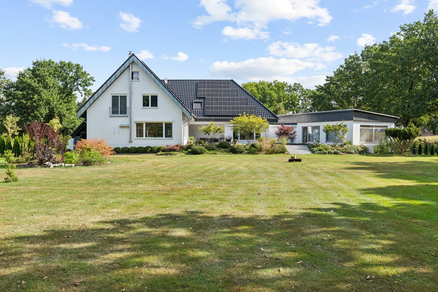 A white house with a black roof and solar panels sits on a large green lawn under a blue sky.
