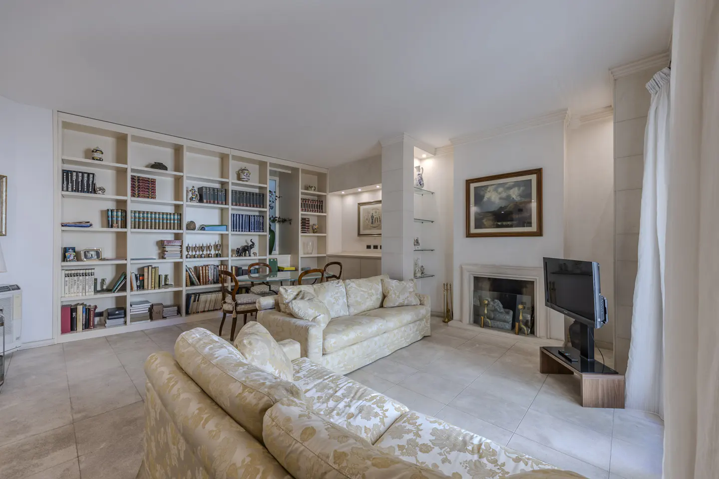 Bright living room with floral sofas, a large white bookcase, a fireplace, and a TV.