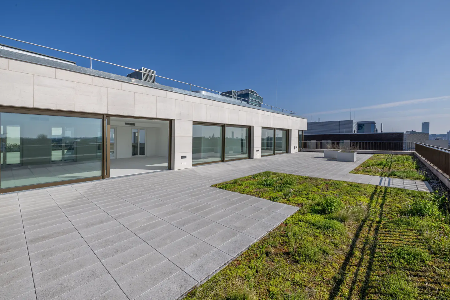 Rooftop patio with gray pavers, green vegetation, and sliding glass doors under a clear blue sky.
