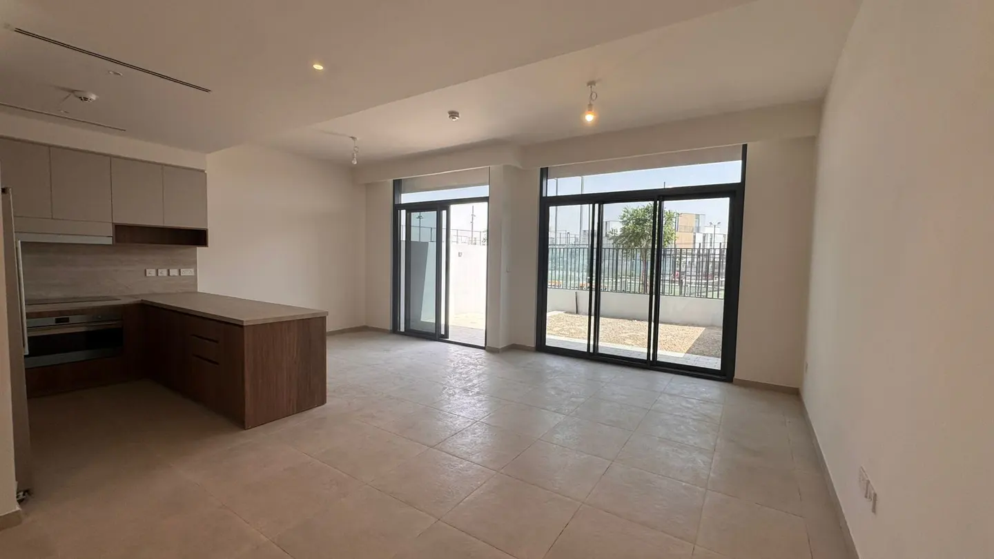 A bright, empty room with tile floors, a kitchen area with brown cabinets, and sliding glass doors to a patio.