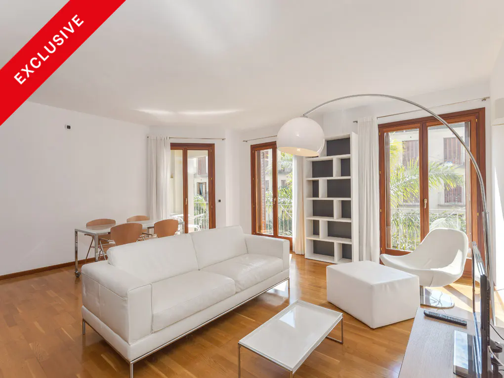 Bright living room with white sofa, chair, and table. Hardwood floors, large windows, and a red "Exclusive" banner in the corner.