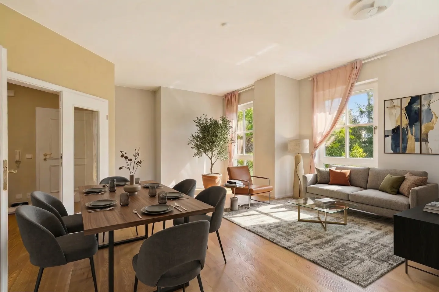 A bright, open-concept living and dining area with wood floors, a dining table with gray chairs, and a gray sofa. Large windows with pink curtains let in natural light.