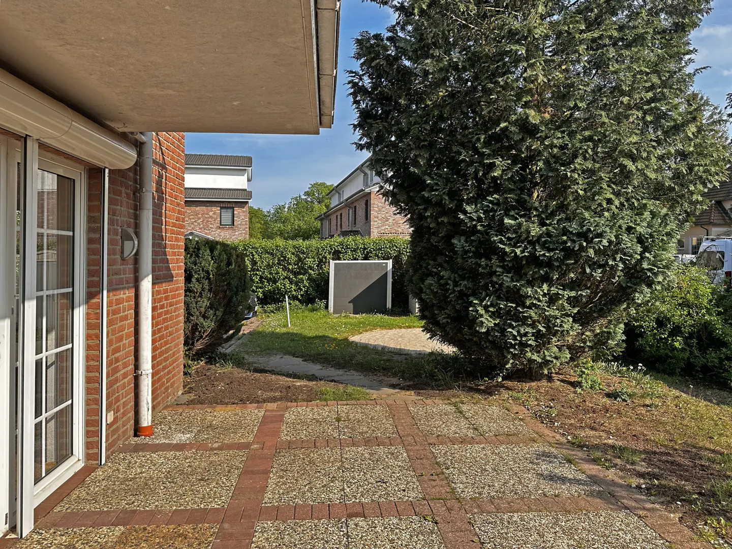 A patio with brick and stone pavers leads to a green lawn with a large evergreen tree. A brick house with white trim is visible.