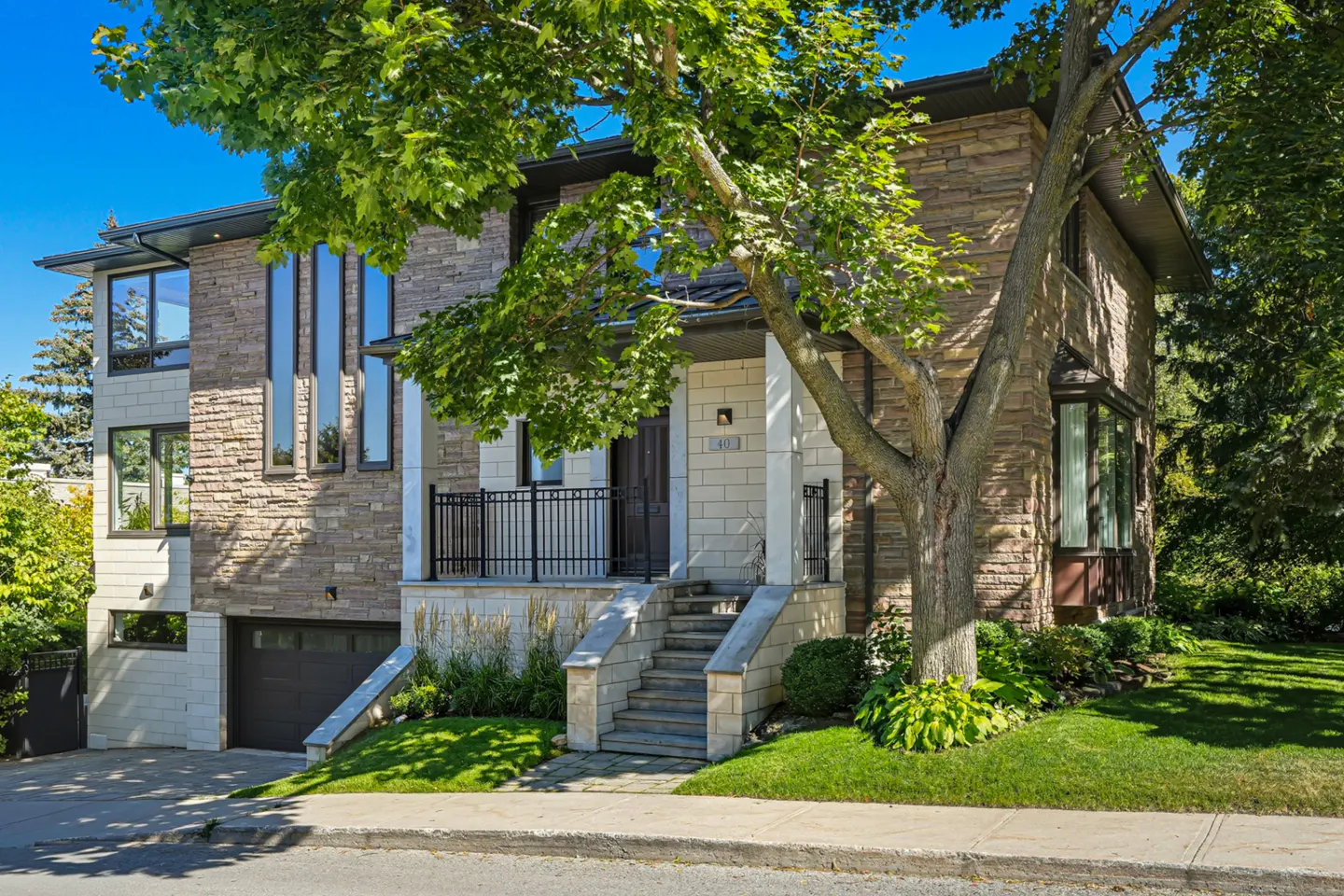 Two-story stone house with a dark roof, black window frames, and a front porch with black railings. A large tree is in the front yard.