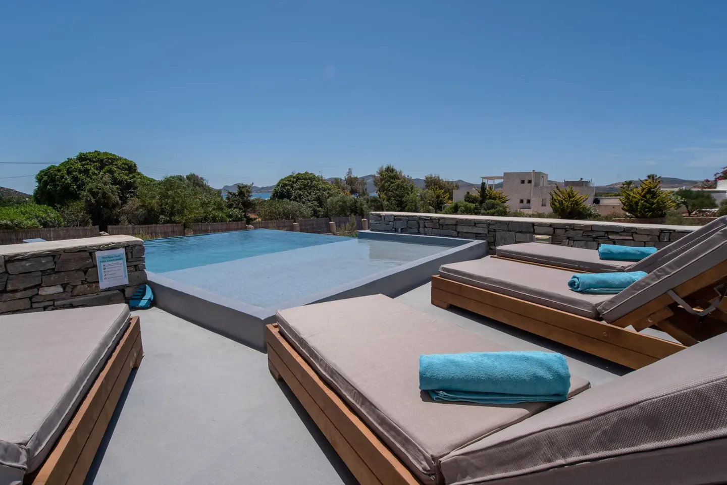 Rooftop pool with lounge chairs and blue towels. Trees and buildings are in the background under a clear blue sky.