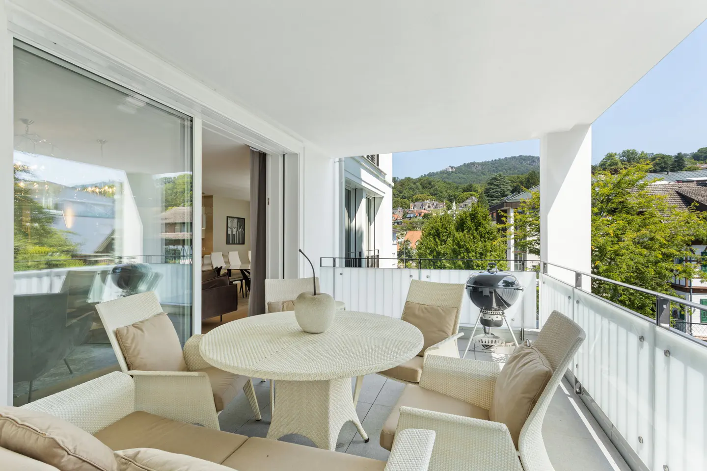 Balcony with white wicker furniture, including a round table, chairs, and a grill. A view of trees and a mountain is visible.