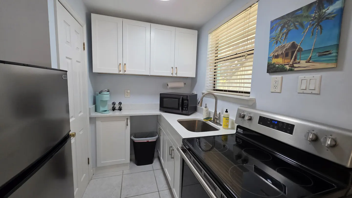 A compact kitchen with white cabinets, stainless steel appliances, and a window with blinds. A tropical painting hangs above the stove.