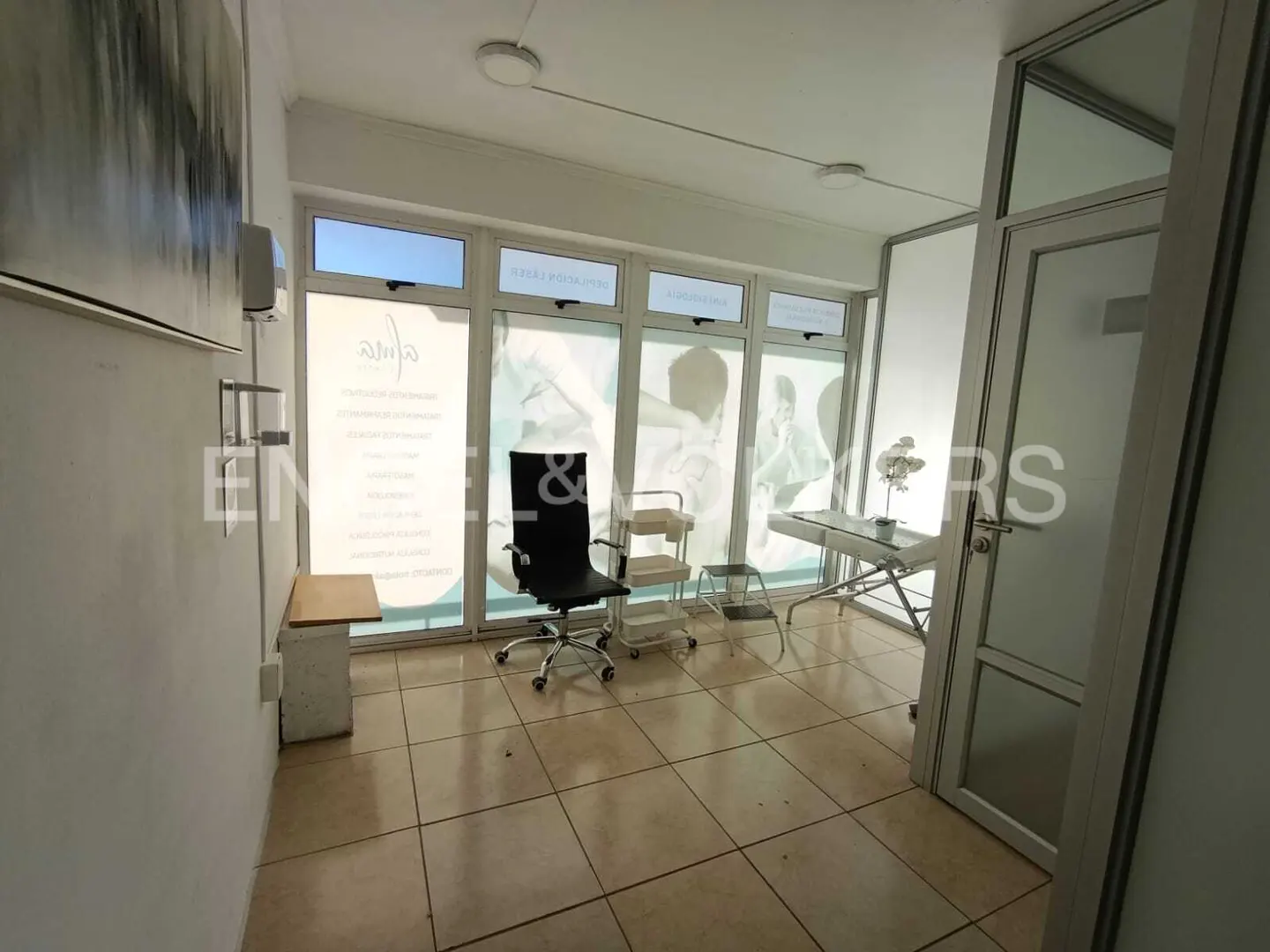 Bright office space with tile floor, black chair, and treatment table near windows with frosted graphics.