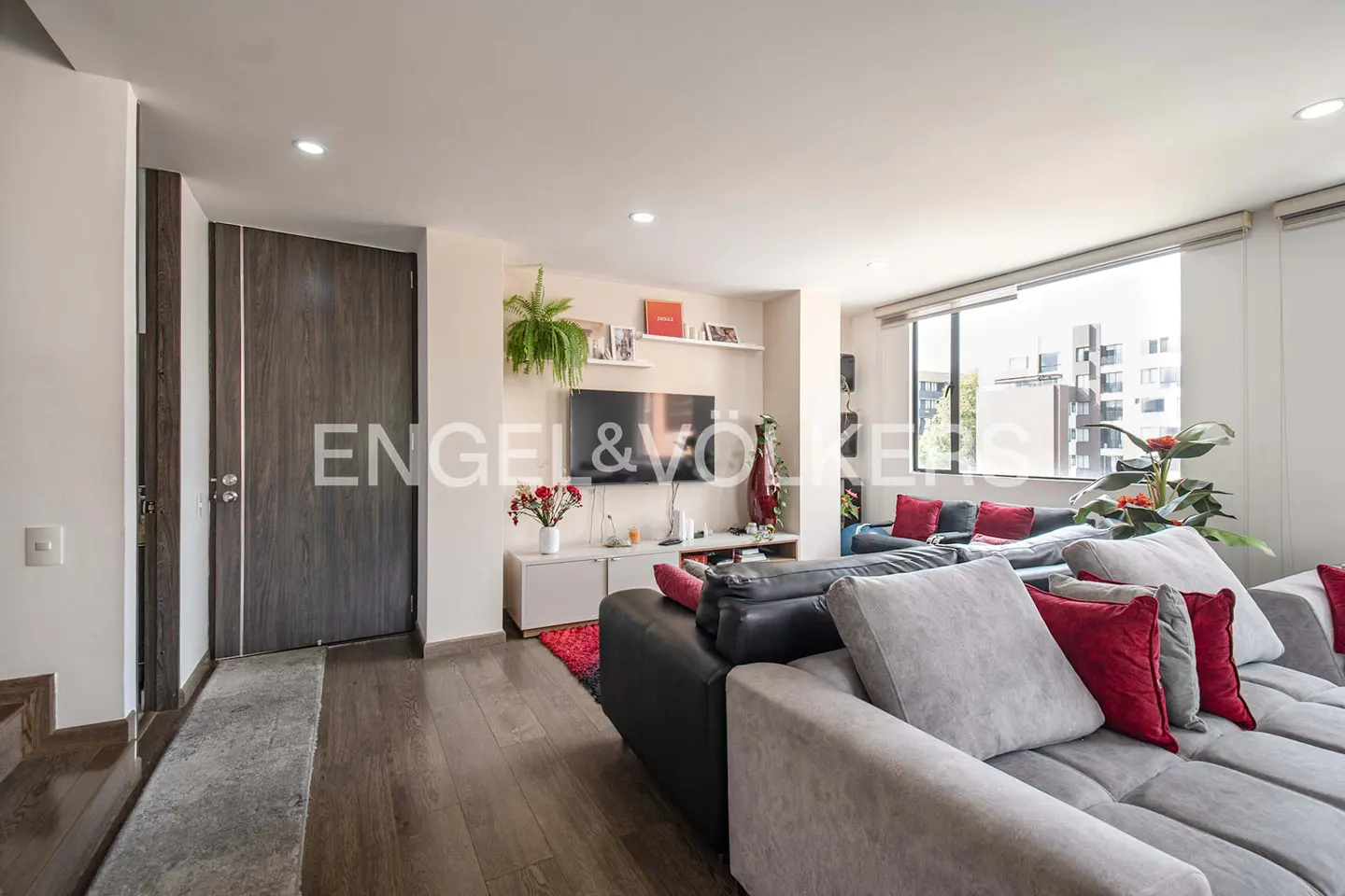 Living room with gray sectional sofa, red pillows, wood floors, TV, and large window with city view.