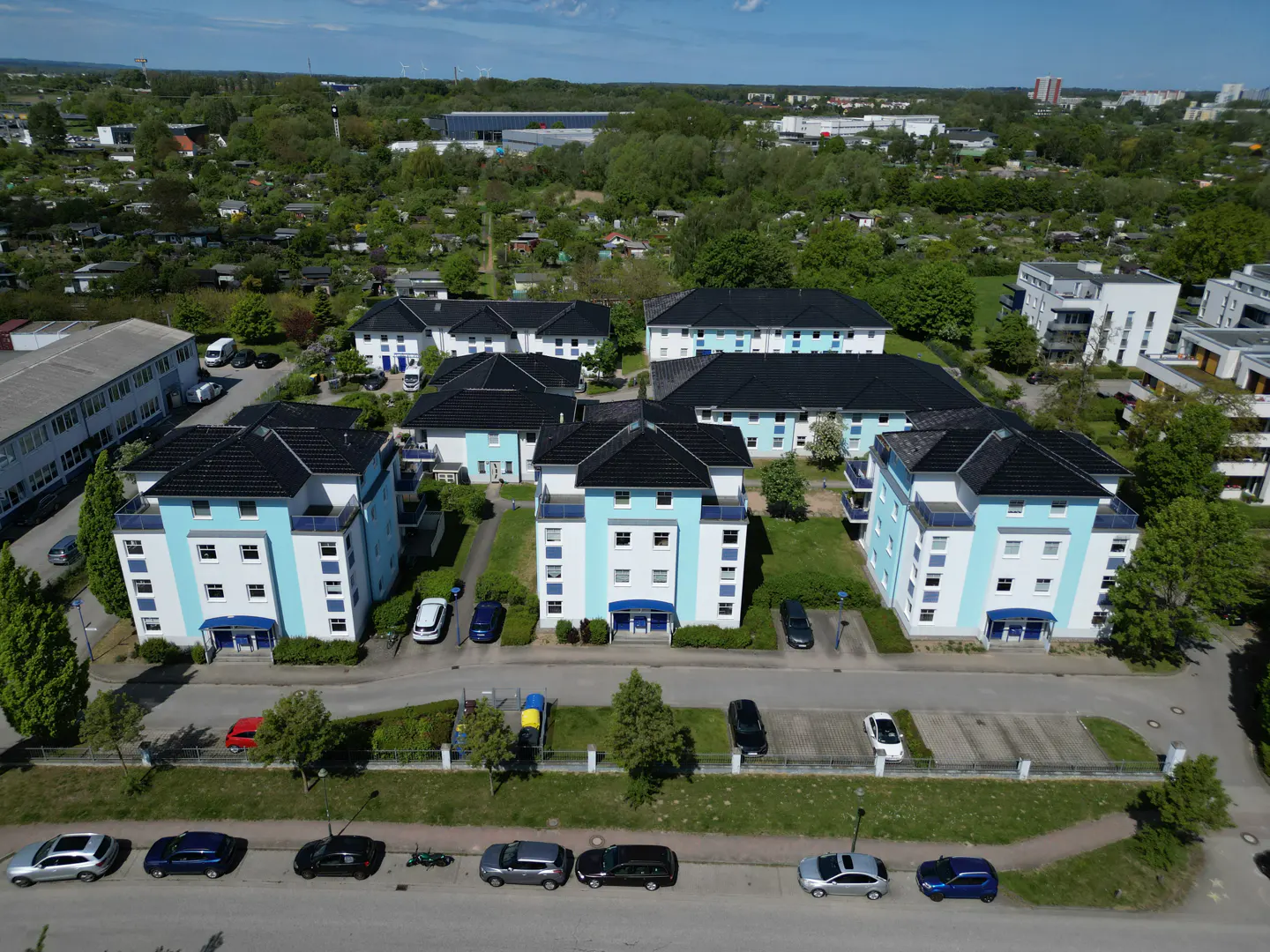 Aerial view of three light blue apartment buildings with dark roofs, surrounded by green trees and parked cars.