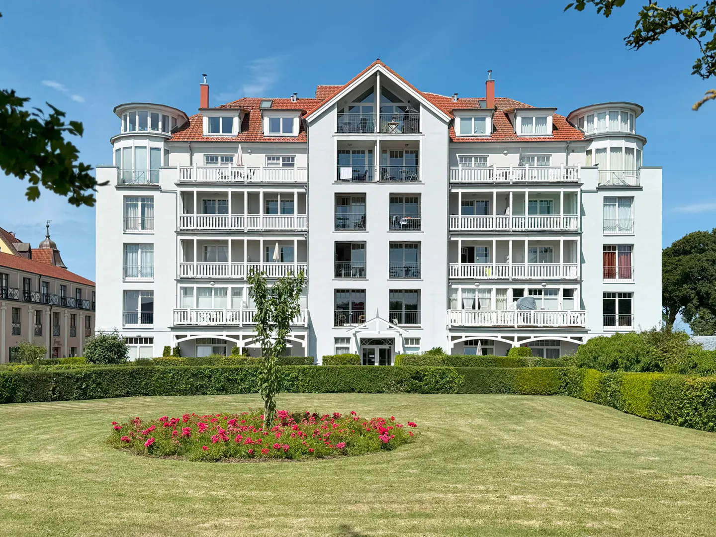 Exterior of a white, multi-story apartment building with balconies and a red tile roof, set against a blue sky. A green lawn and flower bed are in front.