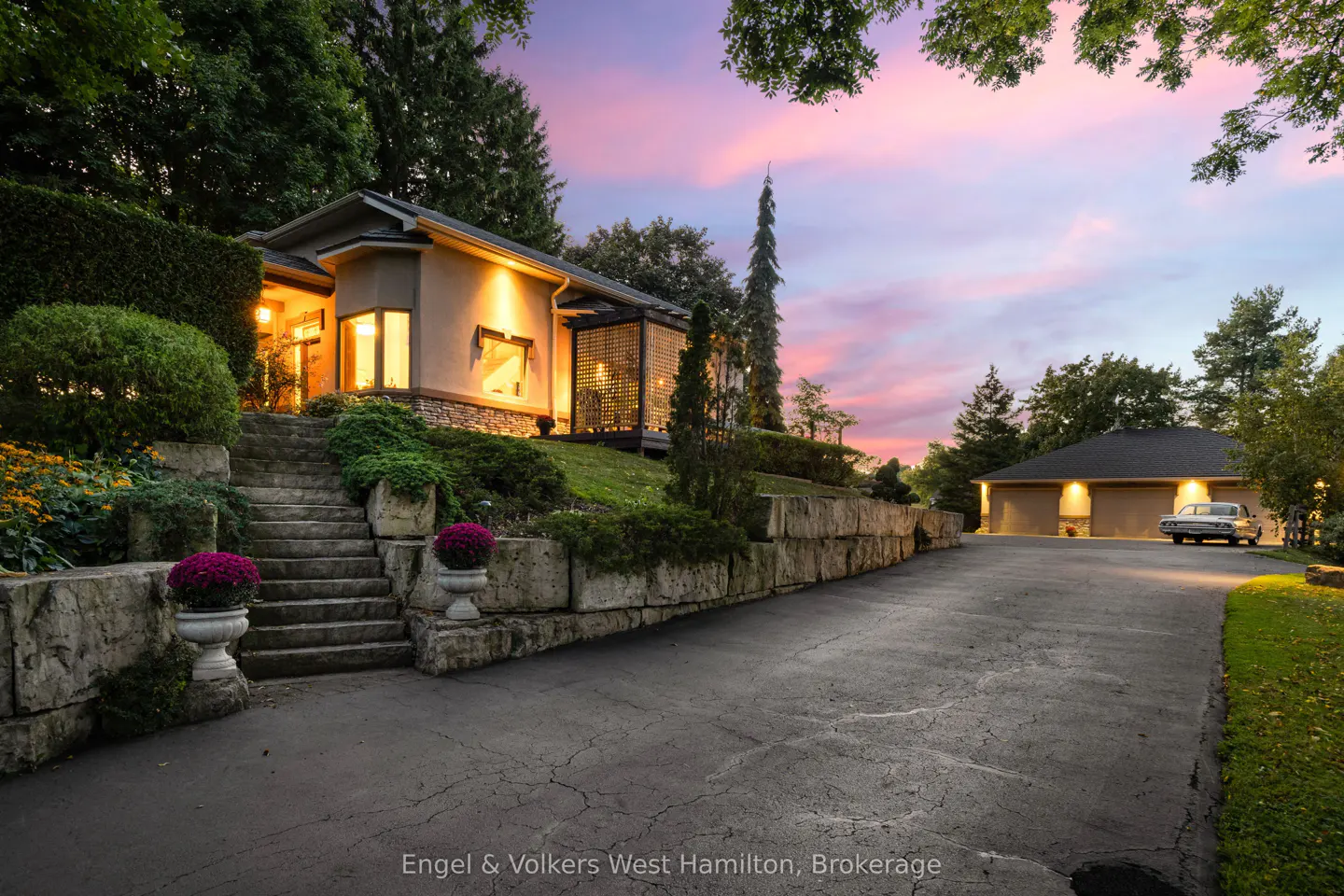 Exterior view of a beige house with stone steps, lush landscaping, and a long driveway leading to a garage with a vintage car at dusk.