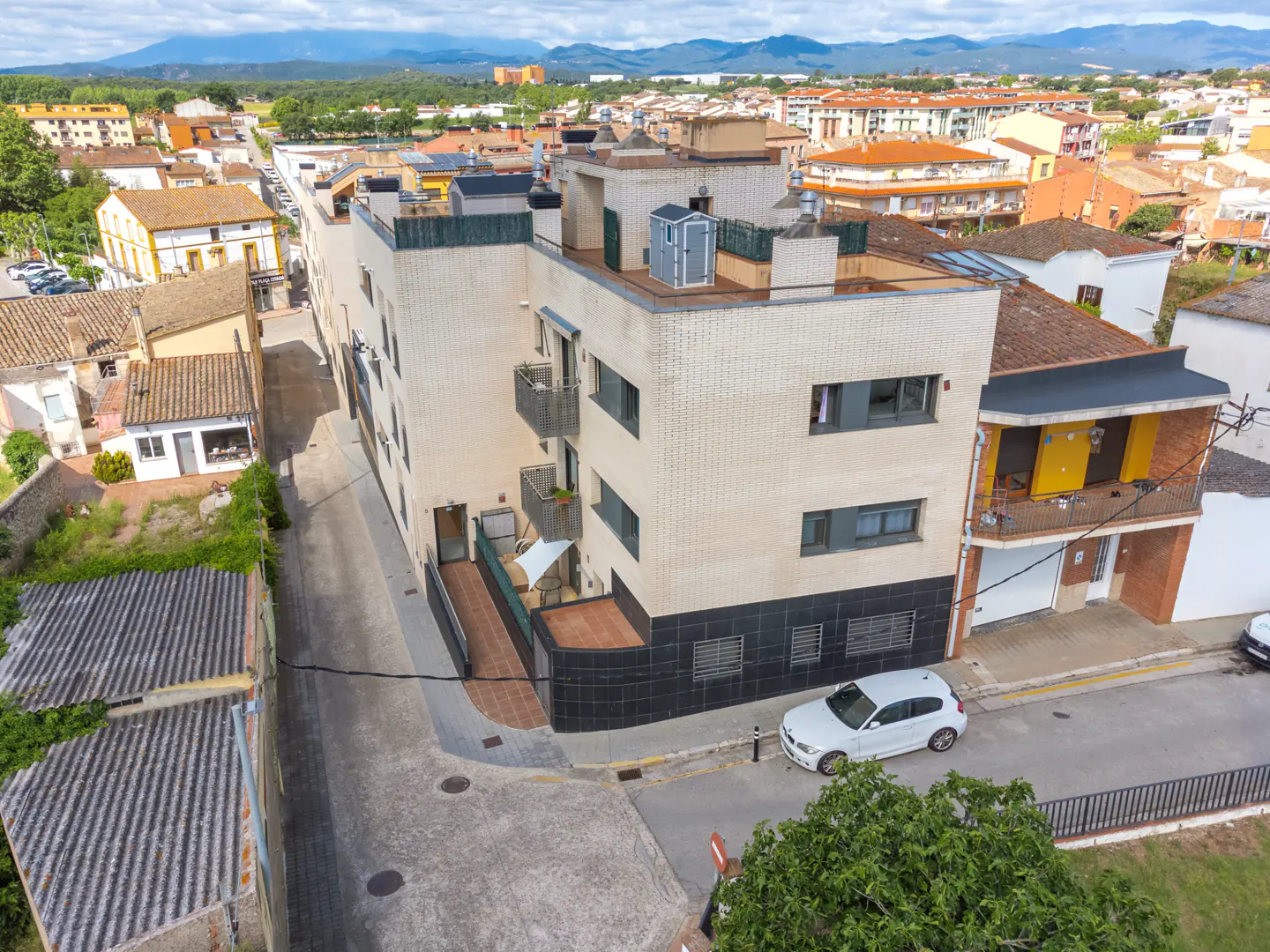 Aerial view of a modern apartment building with balconies, a white car parked nearby, and traditional houses in the background.
