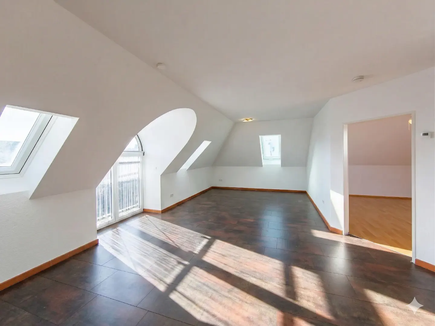 Bright, empty attic room with dark wood floors, white walls, skylights, and a door to another room.