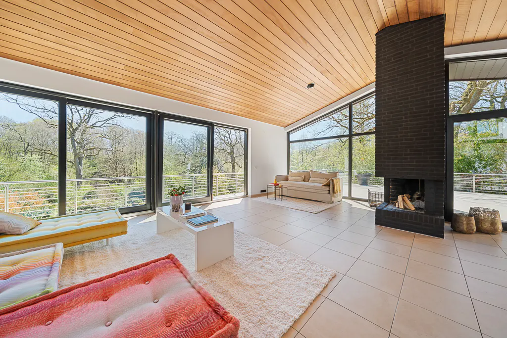 Bright living room with wood ceiling, black brick fireplace, and large windows overlooking trees. Couches and a white coffee table furnish the space.