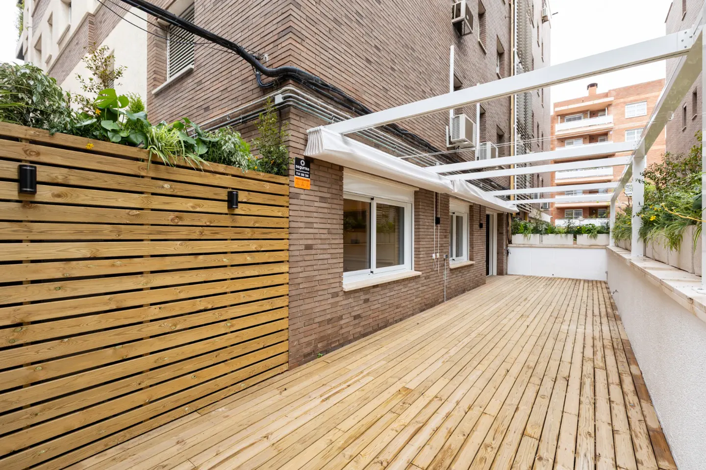 Wooden deck with a white pergola, brick building, and a wooden fence with plants on top.