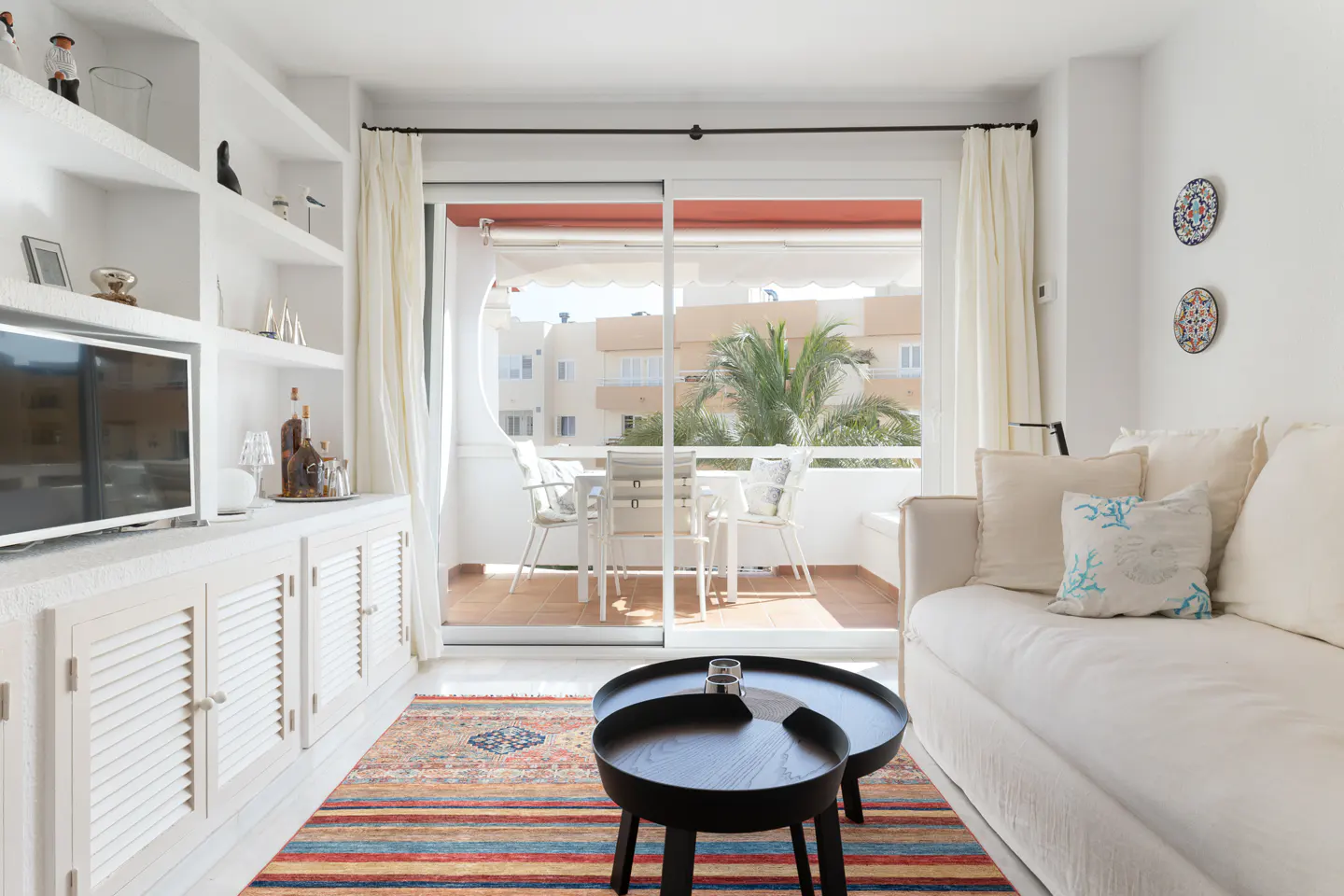 Bright living room with white sofa, black tables, and a colorful rug. Balcony with table and chairs visible through sliding glass doors.