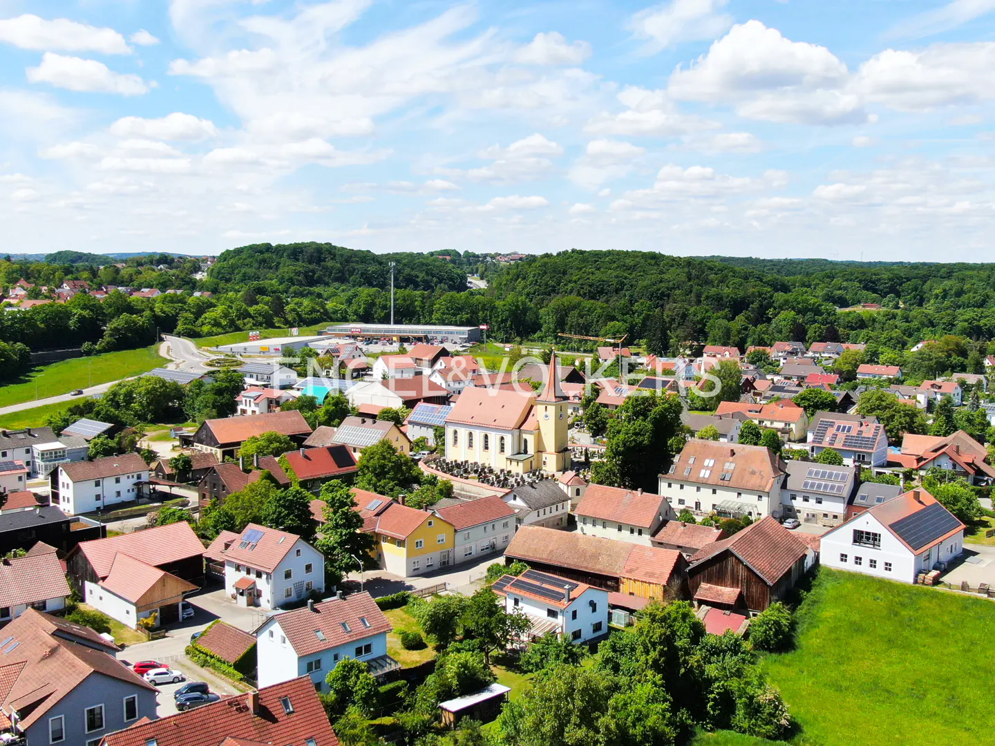 Aerial view of a European village with red-roofed houses, a church with a steeple, and green trees under a blue sky.