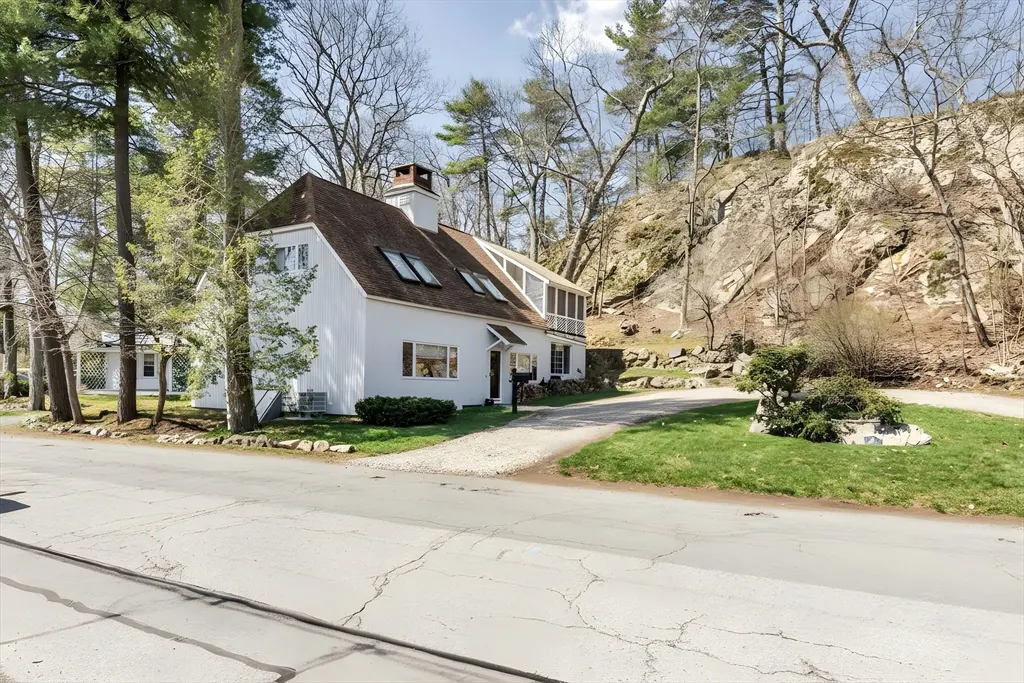 A white house with a brown roof, chimney, and skylights sits near a rocky hill and trees. A gravel driveway leads to the house.