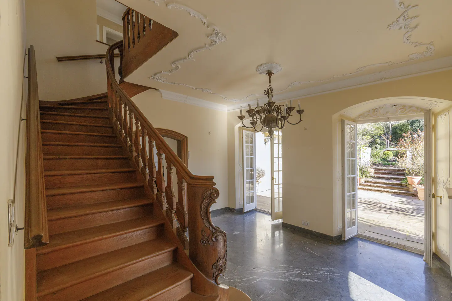 Grand foyer with a winding wood staircase, marble floors, and a chandelier. French doors open to a garden with stone steps.