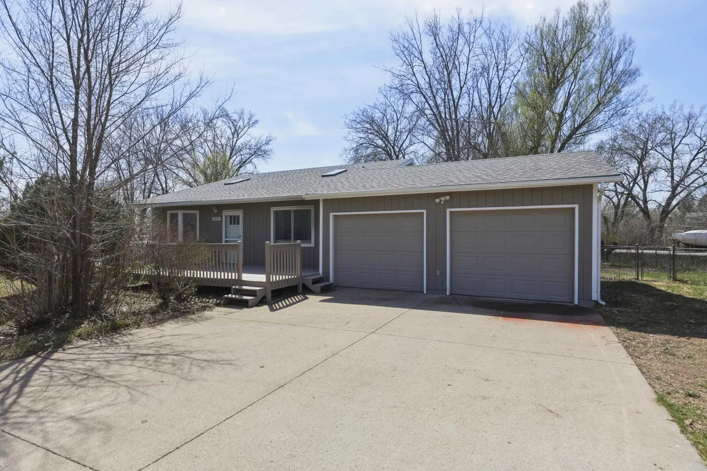 A single-story gray house with a two-car garage and a small wooden porch. A concrete driveway leads to the garage. Trees surround the house.