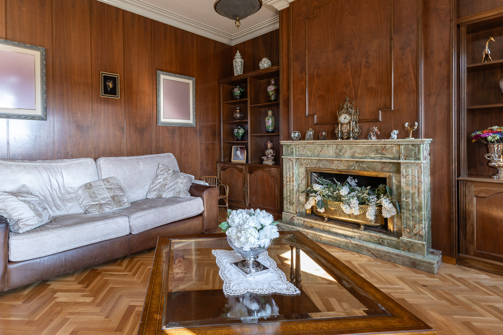 Living room with wood-paneled walls, a white sofa, a glass coffee table with flowers, and a green marble fireplace. [AI-generated text]