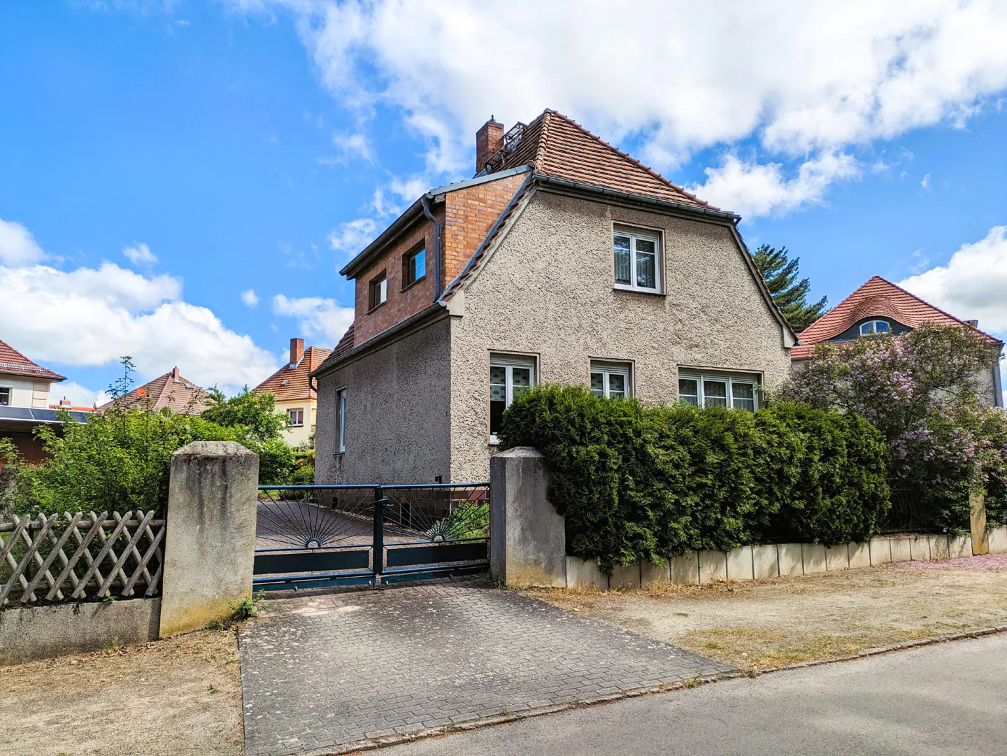 Two-story stucco house with a red-tiled roof, a brick chimney, and a gated driveway under a blue sky.