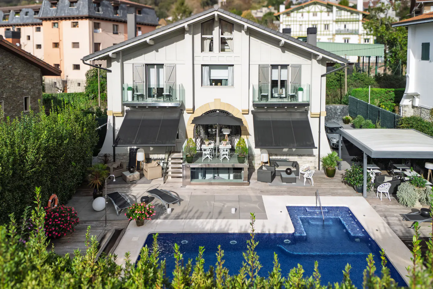 Exterior view of a two-story house with a pool, patio, and balconies. The house is light gray with black awnings and shutters.