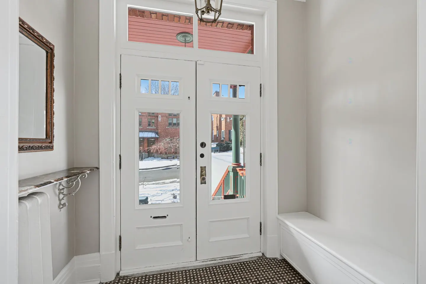 Entryway with white double doors and transom windows. A mirror and marble-topped table are on the left, and a white bench is on the right.
