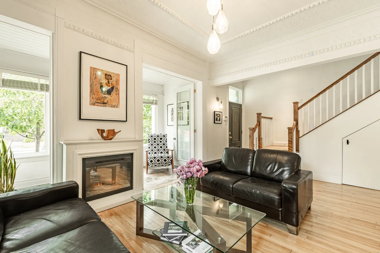 Bright living room with hardwood floors, black leather sofas, glass coffee table with flowers, and a fireplace with art above.