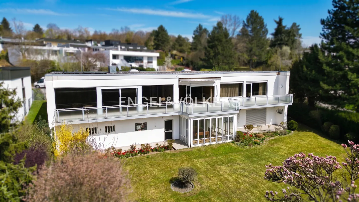 Aerial view of a modern, white house with balconies, a sunroom, and a green lawn. Engel & Völkers logo on the window.