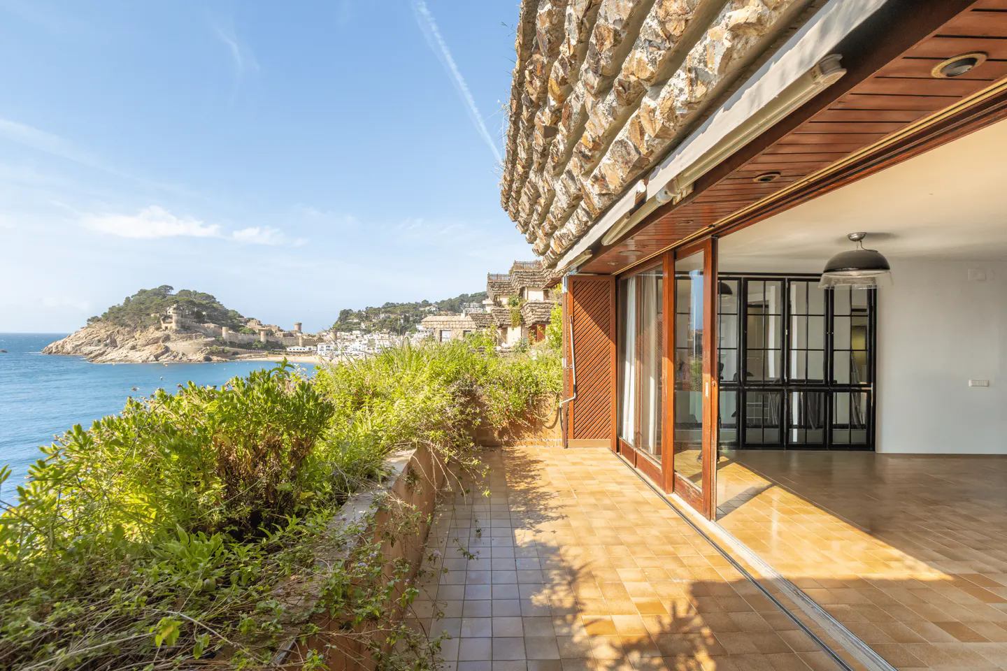 Tiled balcony overlooking the Mediterranean Sea and a coastal town, with large glass doors opening into a modern interior.