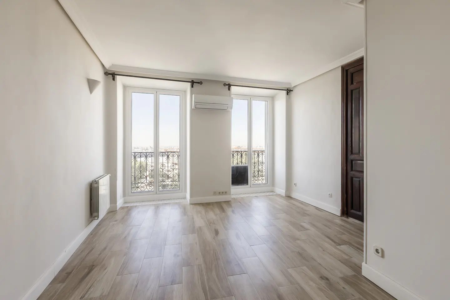 Bright, empty room with wood-look tile floor, white walls, and two windows with black iron balconies. A dark wood door is on the right.