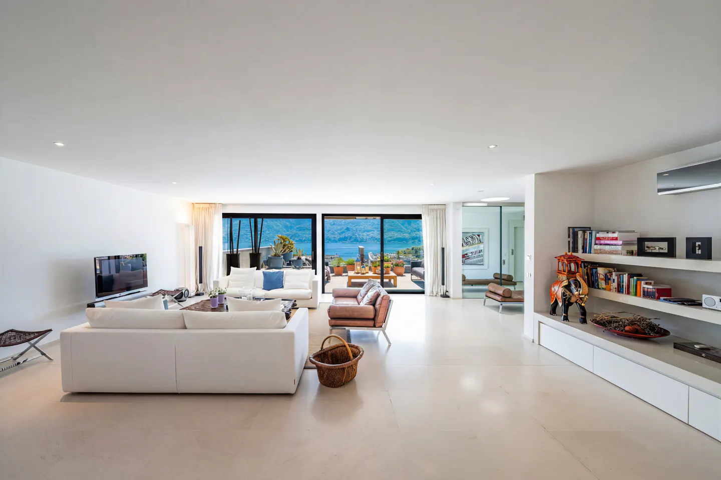 Bright, modern living room with white sofas, a brown leather chair, and a view of the ocean through sliding glass doors. A bookshelf is on the right.