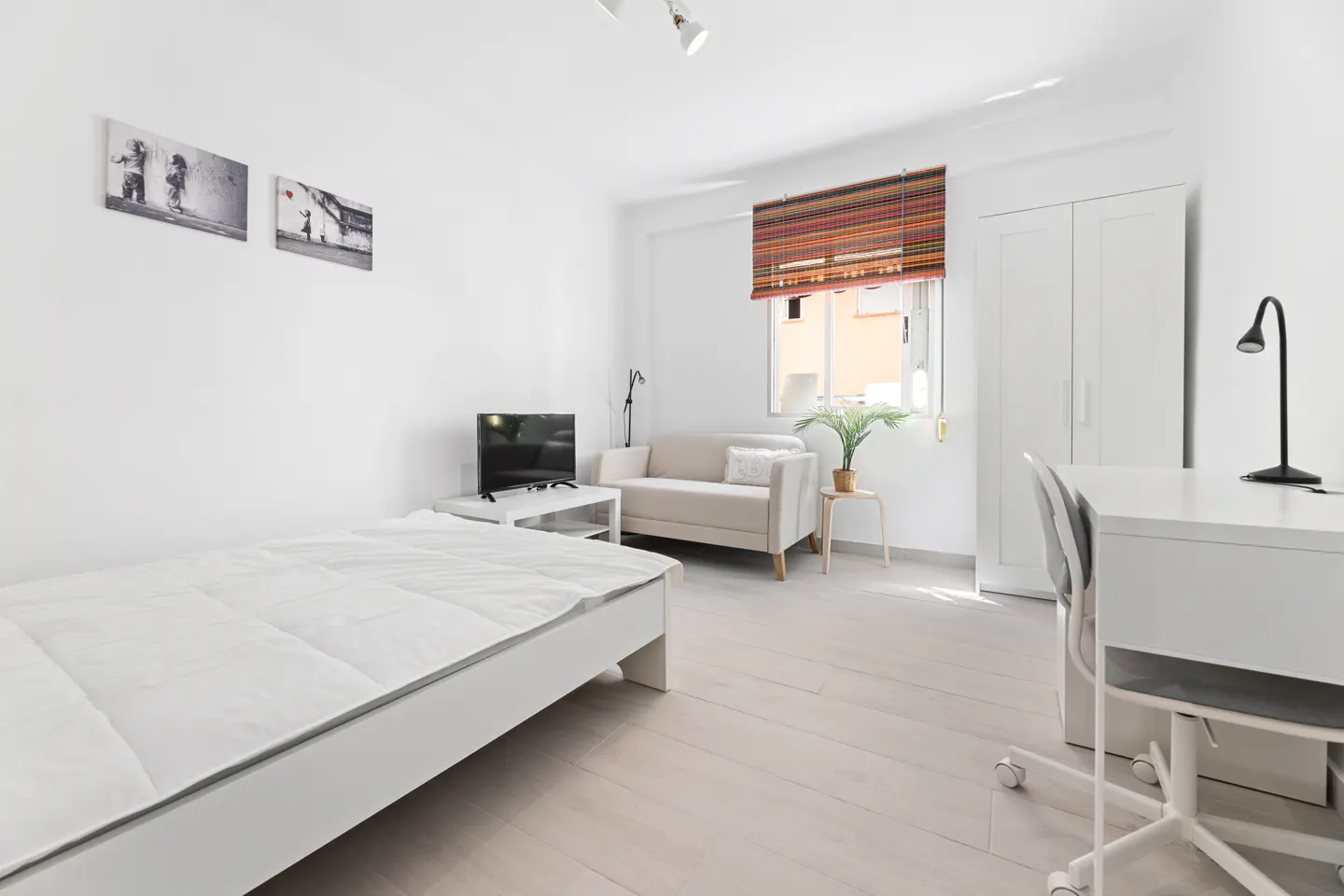 Bright, all-white studio apartment with bed, sofa, desk, and TV. Two framed art prints hang on the wall. A colorful blind covers the window.