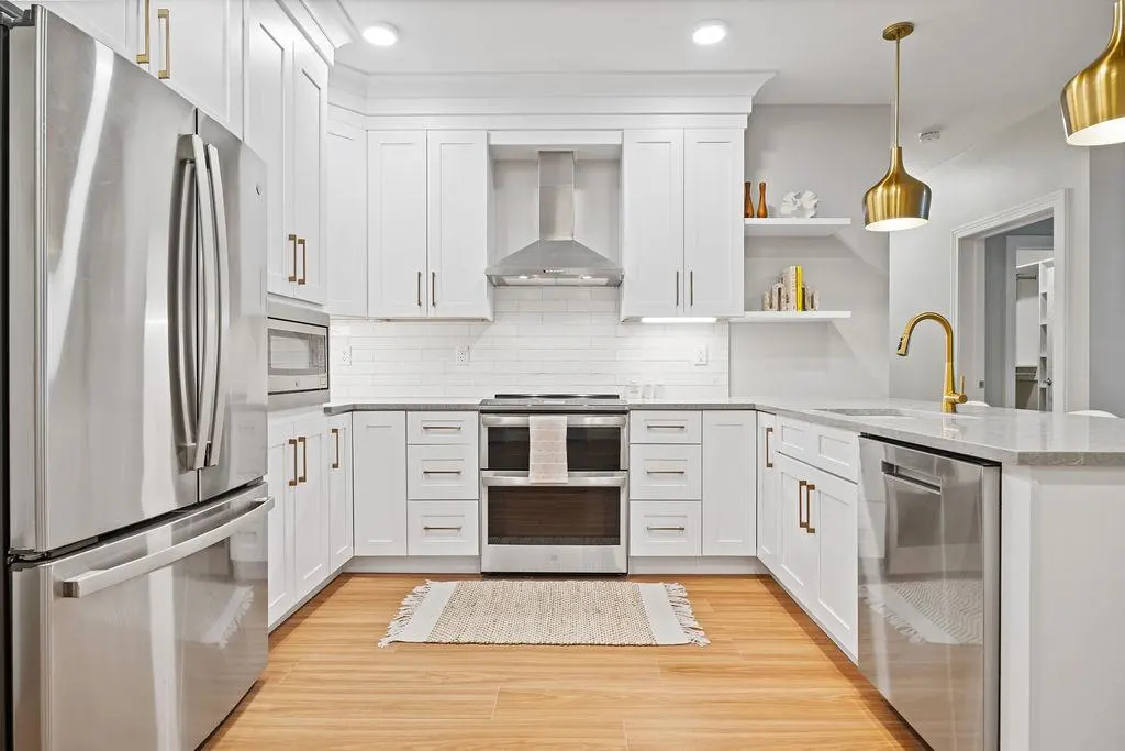 Bright kitchen with white cabinets, stainless steel appliances, and gold accents. Wood floors and a neutral rug add warmth.