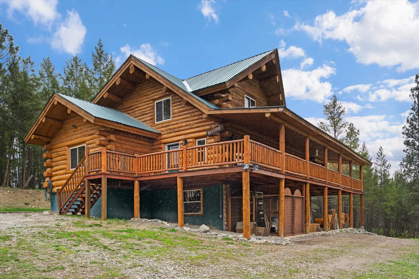 Two-story log cabin with a green metal roof and a large wooden deck, surrounded by trees and a blue sky.