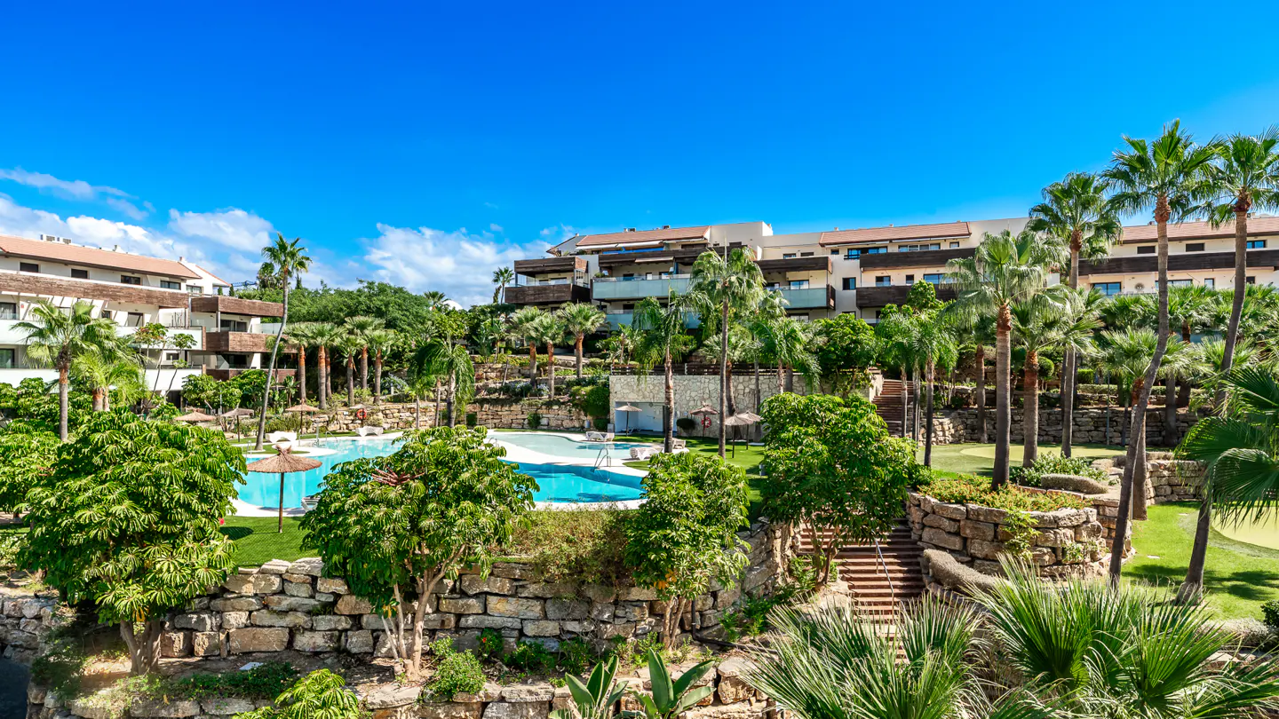 Resort view with a blue pool, palm trees, and stone walls under a clear blue sky.