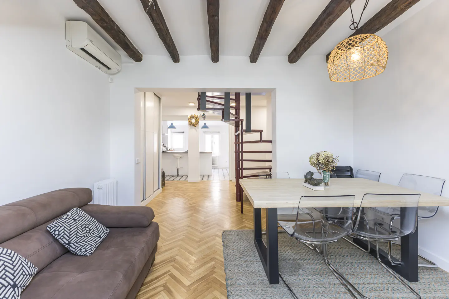 Living room with brown sofa, light wood table with clear chairs, and spiral staircase in the background. Exposed wood beams on the ceiling.