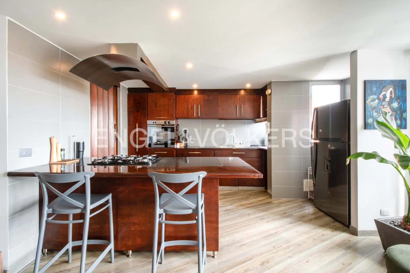 A modern kitchen with wood cabinets, a center island with gray stools, and a black refrigerator.